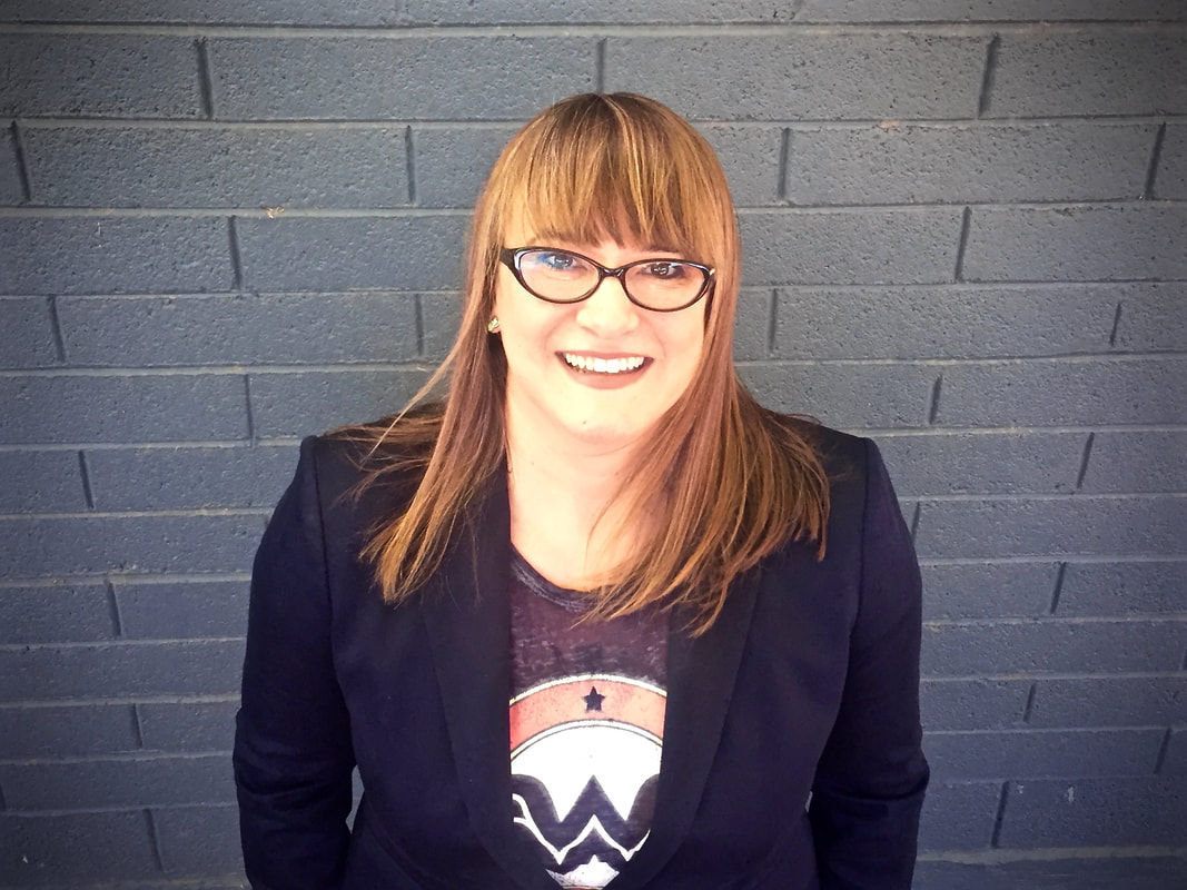 Photograph of Jacqueline Wernimont. She is slightly facing up, toward the camera. Behind her, there is a gray brick wall. She has medium-length, red hair that is down. She is wearing oval, thick-rimmed glasses. She is wearing a black blazer over a t-shirt with a Wonder Woman logo.