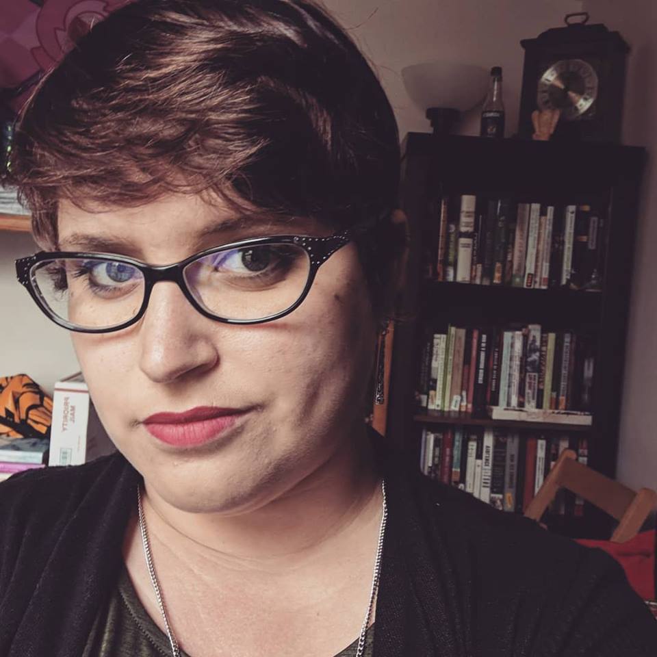Photograph of Gianna May Sanchez. She is slightly facing to the left of the camera, with a small smile. She is wearing oval, thick-rimmed, black glasses and lipstick. She has on a black cardigan and a silver necklace. Behind her, there is a bookcase filled with various books and general clutter. Her brown hair is pixie-cut short and is swooped to the right.