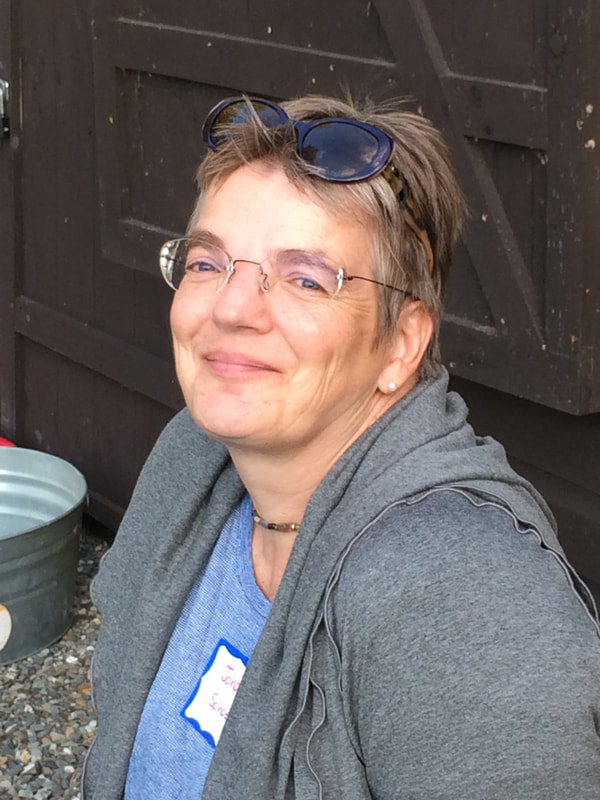 Picture of Johanna Schoen. She is slightly facing up, lookin gat the camera and smiling. She has a brown door background behind her. She is wearing a pair of oval, light-frammed glasses and has a pair of dark, tortoise shell sunglasses on her head. She has short, light brown, pixie-cut hair. She is wearing a gray jacket over a light blue top and is wearing silver stud earrings and a brown-beaded choker necklace.