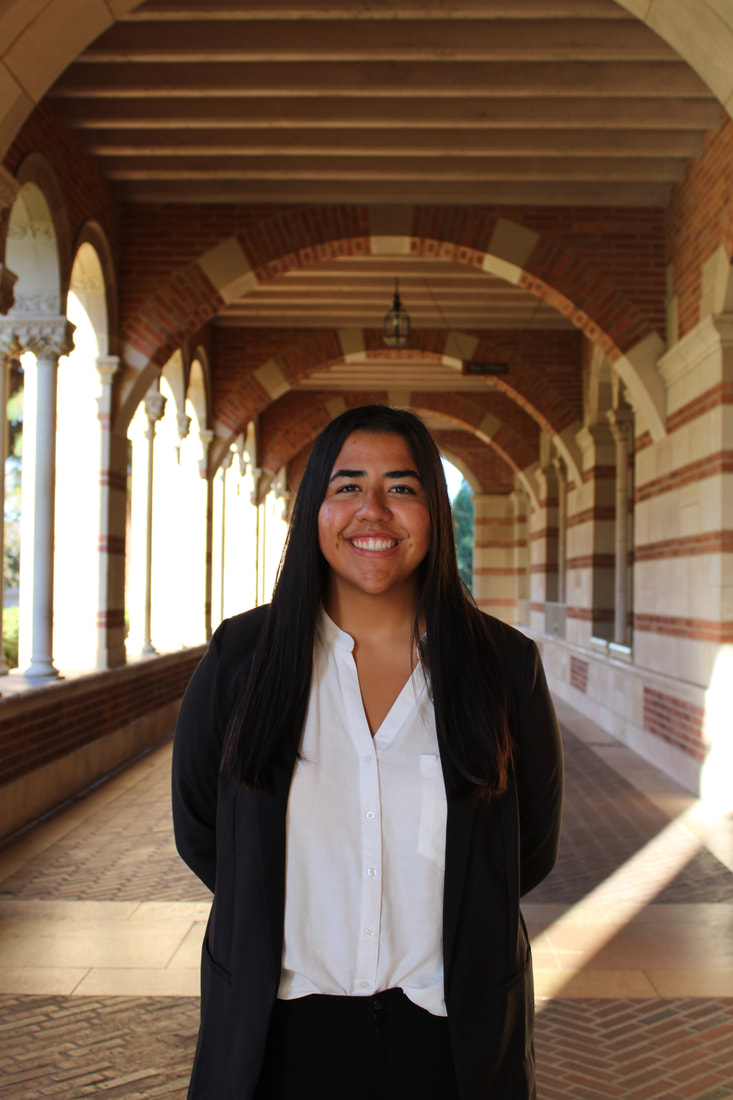 Photograph of Julianna Flores. She is facing the camera, smiling. She is standing in an open, arched walkway with white and red bricks. She is wearing a black blazer over a white, button-up shirt. She has long, black hair that is down.