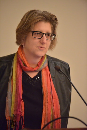 Photograph of Alexandra Minna Stern. She is slightly facing away from the camera, and appears to be giving a talk. She has short, brown hair and is wearing a pair of thick-frammed, rectangle glasses. She is wearing silver earrings and a colorful scarf. She is wearing a black jacket over a black top. A microphone is in the bottom right corner of the shot.